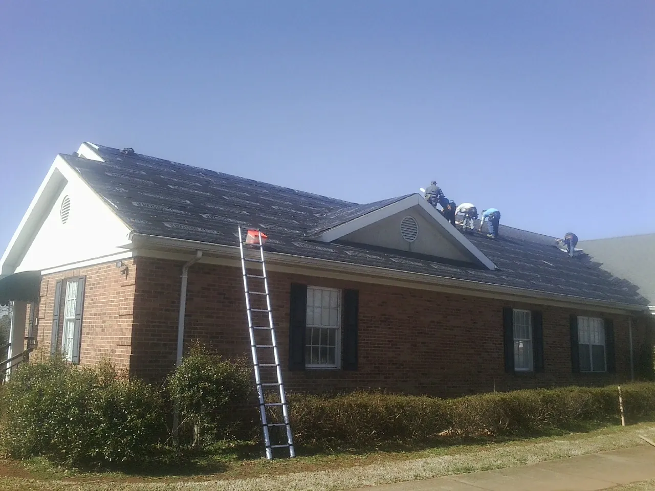 Crew tearing shingles off a brick ranch roof with a ladder set — Emergency Roof Repair work by our crew in Cobb
