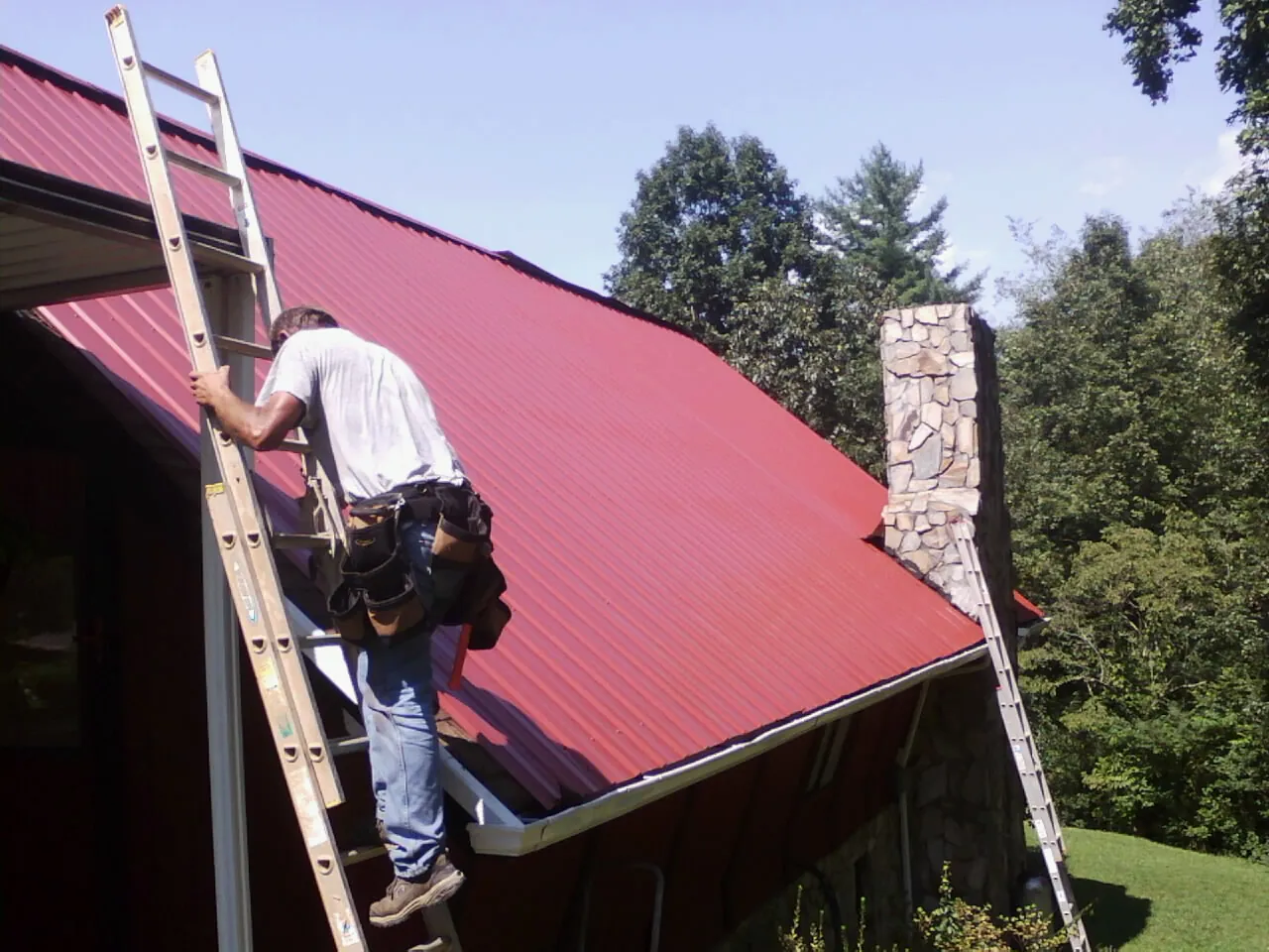 Roofer on a ladder beside a red metal roof and stone chimney — Soffit & Fascia Repair by Roofing Brothers in Cobb